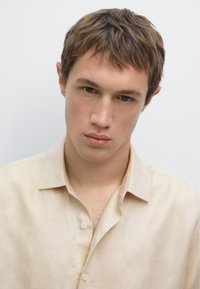 Camicia in lino beige chiaro con colletto a bottoni, caratterizzata da una leggera lucentezza. Il tessuto appare liscio con una vestibilità comoda.