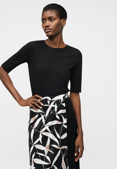 Γυναίκα που φοράει μαύρο εφαρμοστό τοπ με μανίκια μέχρι τους αγκώνες και μαύρη φούστα με λευκά και καφέ φυλλαρά μοτίβα, ποζάροντας με το χέρι στη μέση.