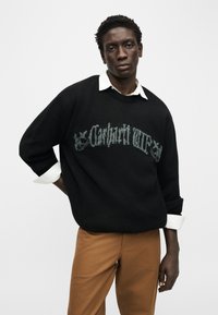 Νέος άνδρας που φοράει μαύρο πουλόβερ με λευκό κείμενο πάνω από λ ευκό πουκάμισο με γιακά και καφέ παντελόνι, στέκεται μπροστά σε απλό φόντο.