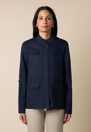 Frau mit langen dunklen Haaren, die goldene Creolen, eine marineblaue Jacke mit vier Fronttaschen und beige Hosen trägt, steht vor einer schlichten beigen Wand.