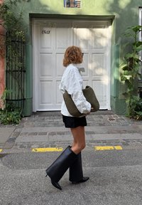 Veste matelassée blanche, shorts noirs, bottes noires hautes avec bouts pointus et un sac tissé vert olive portés en marchant sur une rue en pavés.