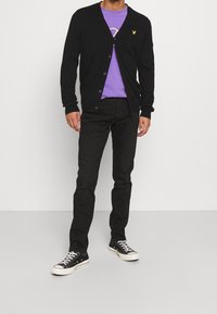 Svart kardigan över en lavendelfärgad t-shirt, i kombination med slim-fit svarta jeans och svarta sneakers. Kardiganen har knappar och en logotypdetalj.