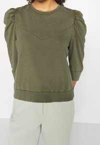 Olivgrön sweatshirt med puffärmar, rund halsringning och ett V-format sömdetalj över bröstet. Mjukt bomullsmaterial.