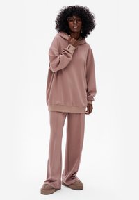 Oversized blushroze hoodie met een textuur in de capuchon, gecombineerd met bijpassende ontspannen broek en lichtbruine instapschoenen.