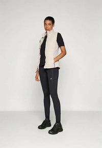Lichtbeige mouwloze vest met een zachte textuur over een aansluitend zwart T-shirt, gecombineerd met donkere leggings en zwarte sportieve schoenen.