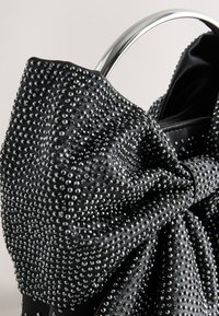 Juoda rankinė su tekstūruotu audiniu, padengtu sidabriniais vinukais, dekoruota dideliu kaspino elementu ir blizgiu metaliniu rankena.