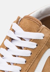 Hnědá semišová teniska s bílými tkaničkami, zdobená bílým bočním pruhem, prošitými akcenty a texturovaným svrškem. Detailní pohled na oblast šněrování.