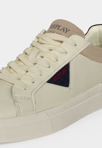 Baskets couleur crème avec détail en daim beige, lacets blancs et un patch bleu marine avec texte rouge "REPLAY" sur le côté.