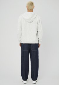 Personne avec des cheveux blonds courts portant un sweat à capuche gris clair, un jean bleu foncé ample et des baskets blanches, se tenant de dos sur un fond uni.