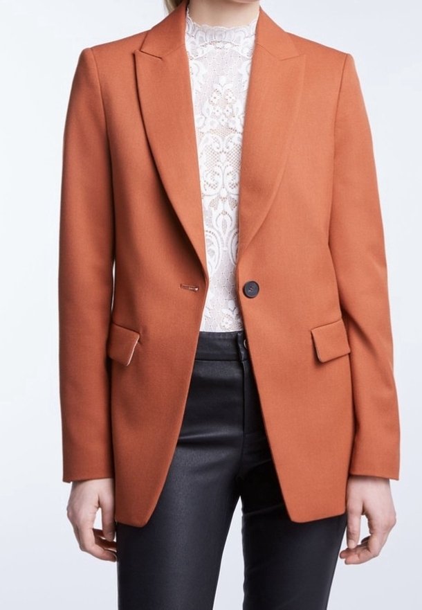 Blazer arancione su misura con rever a punta, bottone singolo, due tasche frontali e una texture liscia, indossato sopra un top in pizzo.