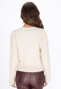 Krém színű kötött pulóver strukturált mintával, bordázott mandzsettával és szegéllyel, valamint fonott kötött részletekkel az ujján. Barna műbőr nadrággal viselve.