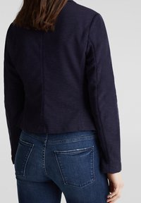 Chaqueta azul marino con un diseño ajustado, tela texturizada y mangas largas. Combinada con jeans ajustados de color azul oscuro con bolsillos traseros.