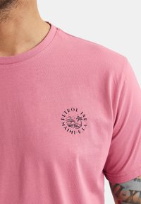 Rosa Baumwoll-T-Shirt mit einem runden Logo-Druck auf der linken Brust, das Text sowie eine Grafik von einer Palme und einer Welle zeigt.