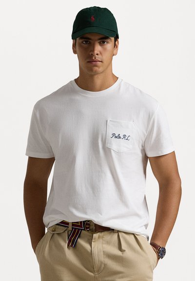 White cotton t-shirt with a chest pocket featuring blue "Polo R.L." embroidery, paired with beige trousers and a dark green cap.