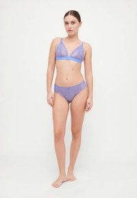 Donna in piedi a piedi nudi che indossa un set coordinato di reggiseno e mutandine in pizzo trasparente azzurro chiaro contro uno sfondo semplice chiaro.