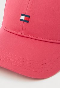 Rožinė beisbolo kepuraitė, pagaminta iš audinio su raižyta tekstūra. Priekyje yra mažas siuvinėtas logotipas mėlynos, baltos ir raudonos spalvų.