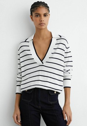 Žena s vrkočmi, oblečená v bielom svetri s hlbokým výstrihom do V a čiernymi horizontálnymi pruhmi, a tmavých džínsach s vysokým pásom, stojaca pri bielej stene.