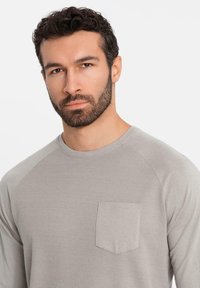 Camisa de manga larga gris claro con textura de canalé y un bolsillo de parche en el lado izquierdo del pecho; cuenta con un cuello redondo clásico y mangas raglán.