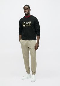 Melns džemperis ar zeltītu EAT logotipu, bēšas šauras bikses un baltas kedas. Modelis demonstrē brīvu piegriezumu un ikdienišķu stilu uz vienkārša fona.