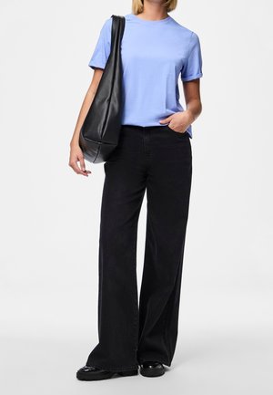 Osoba ubrana w jasnoniebieską koszulę z krótkim rękawem, czarne spodnie o szerokich nogawkach, czarne buty oraz niosąca dużą czarną torbę na ramię.