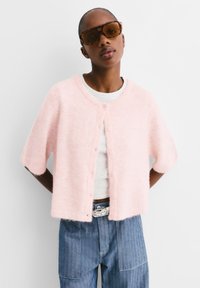 Cardigan rose à manches courtes avec une texture douce et duveteuse, doté d'un col rond et d'une fermeture à boutons, porté sur un t-shirt blanc et un jeans bleu.