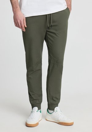 Uomo che indossa pantaloni jogger verde oliva con polsini elastici, sneaker bianche con suola in gomma e una camicia bianca, in piedi con le mani nelle tasche.