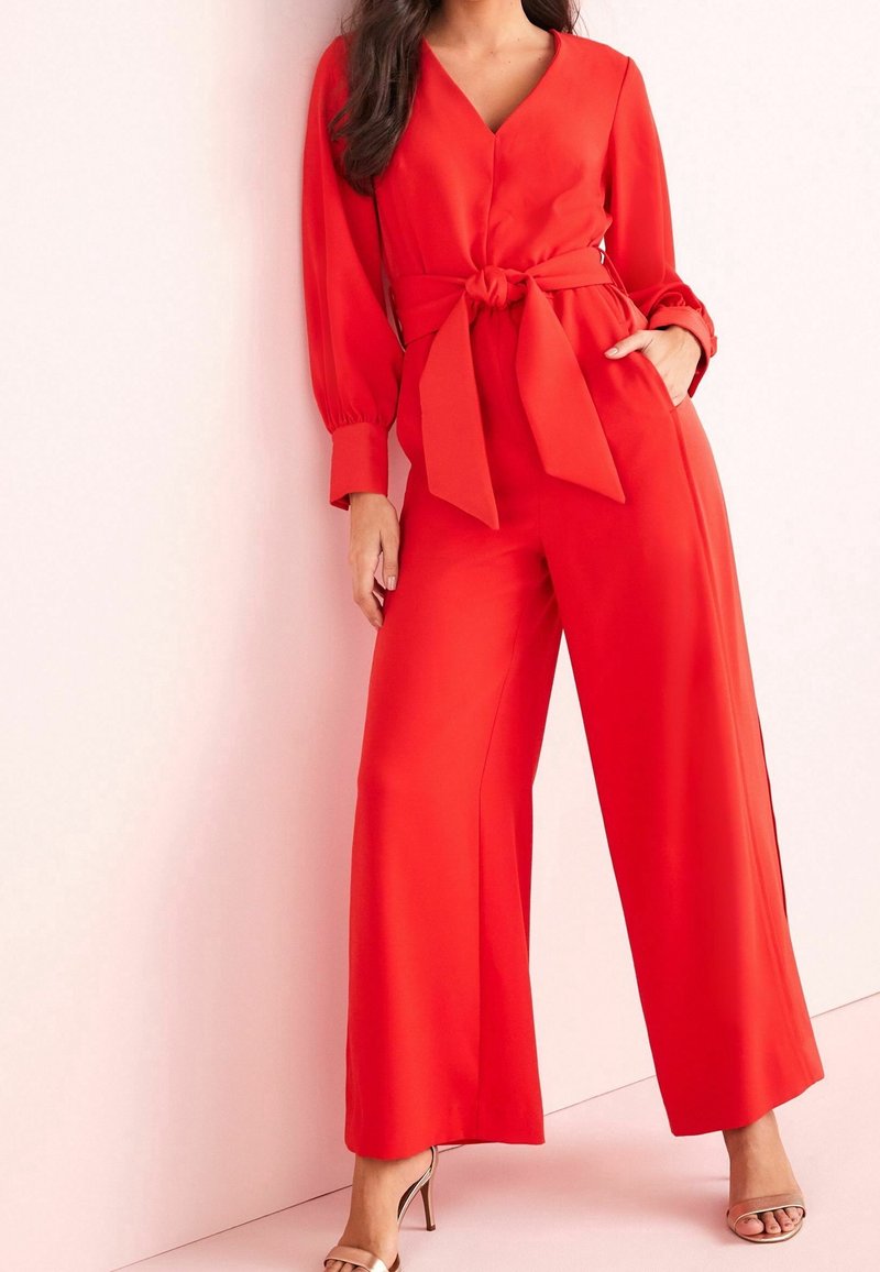 Femme portant une combinaison rouge vif à manches longues avec une ceinture nouée à la taille et un pantalon à jambes larges, debout devant un mur rose clair.