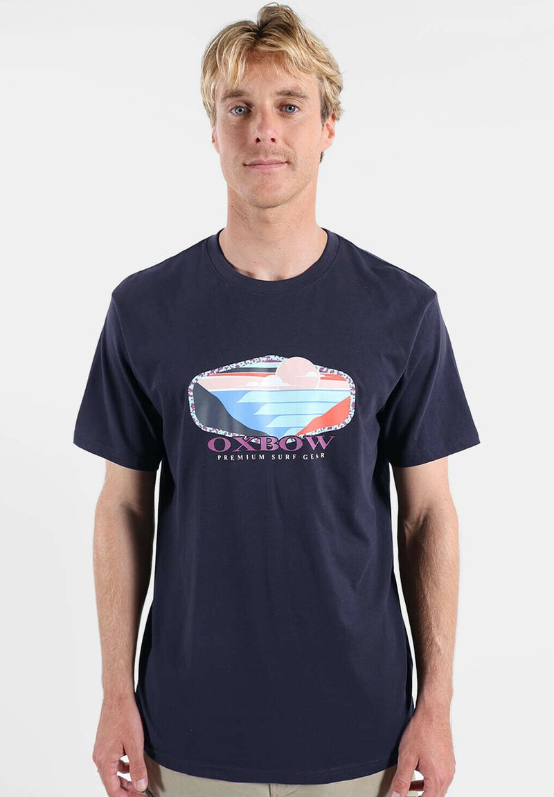 T-shirt en coton bleu marine à manches courtes, avec un graphique coloré représentant des vagues et un soleil, accompagné du texte « Oxbow Premium Surf Gear » sous l'image.