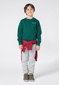 Felpa verde con logo ricamato, pantaloni da tuta grigi e sneakers olive. Maglione rosso annodato in vita. Bambino in piedi, sorridente.