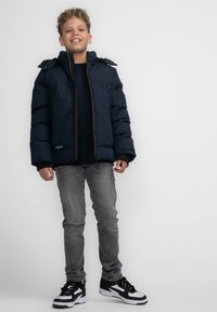 Marineblauer Puffy-Jacke mit Kapuze, ausgestattet mit einem frontalen Reißverschluss und kontrastierenden braunen Nähten. Getragen mit einem schwarzen Shirt, grauen Jeans und schwarzen Sneakern.