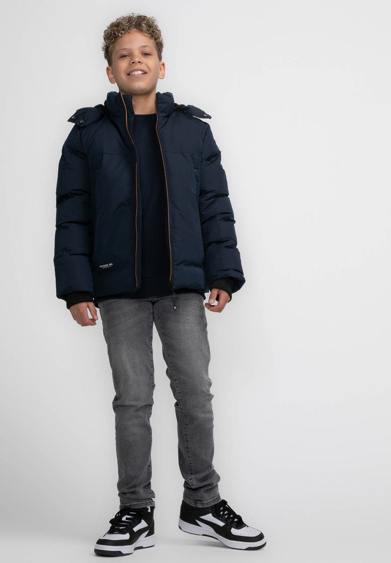Marineblauer Puffy-Jacke mit Kapuze, ausgestattet mit einem frontalen Reißverschluss und kontrastierenden braunen Nähten. Getragen mit einem schwarzen Shirt, grauen Jeans und schwarzen Sneakern.