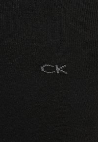 Melnas zeķes ar ribotu tekstūru un mazu baltu "CK" logotipu izšūtu tuvu augšai. Materiāls izskatās mīksts un elastīgs.