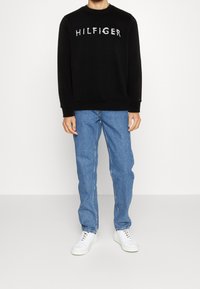 Černá mikina s nápisem "HILFIGER", kulatým výstřihem a žebrovanými manžetami, nošená s modrými džíny a bílými teniskami. Minimalistický design.