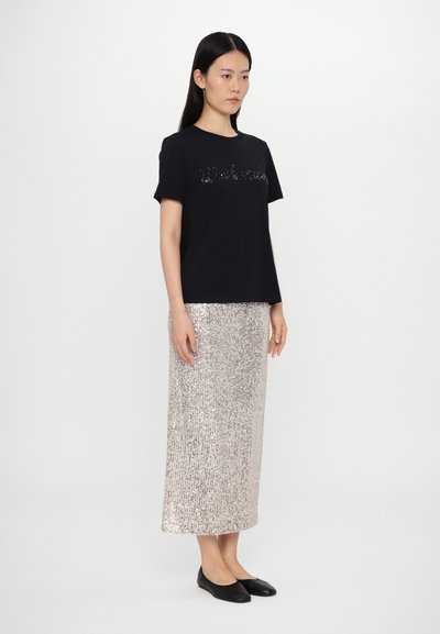 Sort t-shirt med palietteret tekst, parret med en lang, sølv palietskørt. Modellen bærer sorte flade sko og står foran en ensfarvet baggrund.