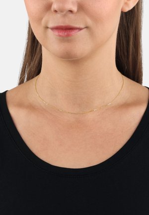 Collana choker sottile e color oro con un design delicato, caratterizzata da piccoli accenti rotondi, indossata contro il colletto di una maglietta nera.