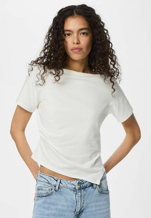 Jeune femme aux cheveux bouclés portant un haut blanc uni à manches courtes et un jean denim bleu clair, debout les mains sur les hanches.