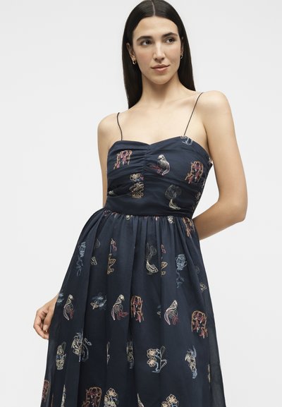 Femme portant une robe bleu marine à fines bretelles avec des motifs abstraits multicolores, debout sur un fond blanc uni.