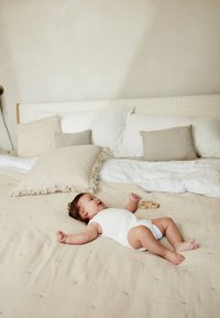 Un copil îmbrăcat într-un body alb zace pe o pătură bej texturată, înconjurat de perne moi în nuanțe neutre și o jucărie din lemn.