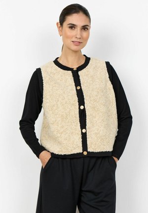 Kvinde iført en beige tekstureret knappet vest over en sort langærmet bluse og sorte bukser, stående med hænderne i lommerne.