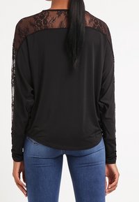Svart långärmad topp med en genomskinlig spetspanel vid axlarna och ärmarna, mjukt tyg och en avslappnad passform, tillsammans med blå jeans.