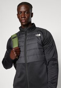 Chaqueta negra y gris con cremallera, sección superior acolchada, ajuste ajustado, cuello alto y logo en el pecho. Cuenta con una correa verde sobre el hombro.