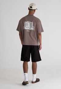 Brown graphic t-shirt featuring a white building illustration on the back, paired with black shorts, white socks, and brown slip-on sandals.