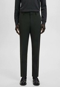 Mężczyzna w ciemnozielonych, dopasowanych spodniach, czarnych skórzanych mokasynach oraz ciemnym swetrze z długim rękawem na jasnym, jednolitym tle.