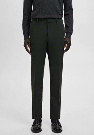 Mężczyzna w ciemnozielonych, dopasowanych spodniach, czarnych skórzanych mokasynach oraz ciemnym swetrze z długim rękawem na jasnym, jednolitym tle.