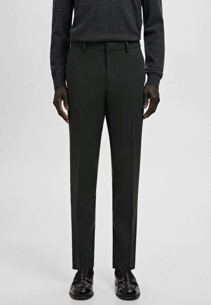 Mężczyzna w ciemnozielonych, dopasowanych spodniach, czarnych skórzanych mokasynach oraz ciemnym swetrze z długim rękawem na jasnym, jednolitym tle.