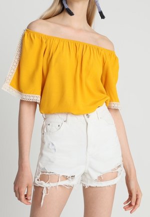 Frau trägt ein gelbes schulterfreies Oberteil mit Spitzenbesatz und hochtaillierte weiße, verwaschene Denim-Shorts.