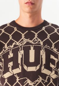 Close-up of a person wearing a brown sweater with beige chain-link pattern and large block letters spelling "HUG" on the chest.