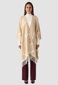 Poncho beige con grandi lettere bianche, apertura frontale e orlo sfrangiato. Indossato sopra un dolcevita bianco e pantaloni bordeaux scuro. Tessuto dalla consistenza liscia.
