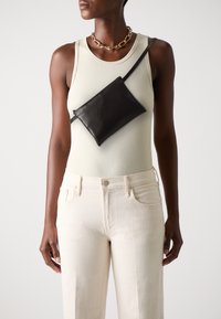 Svart skinn crossover-veske med rektangulær form og glidelås, båret diagonalt over en lysfarget ribbet tanktop og beige jeans.