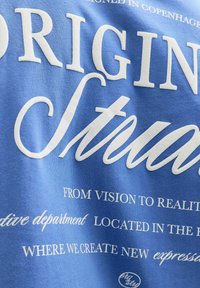 Blauwe katoenen t-shirt met grote witte tekst met verschillende zinnen, waaronder "ORIGINEEL" en "CREATIEVE AFDELING." Pretty textuur.